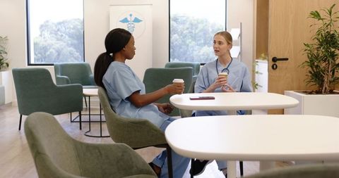 Two nurses sharing coffee and conversing in bright hospital staff lounge during break