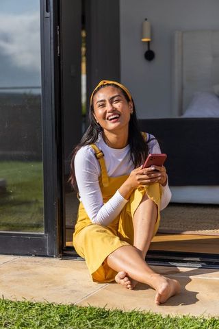 Sunlit asian woman laughing on patio threshold holding smartphone in mustard pinafore