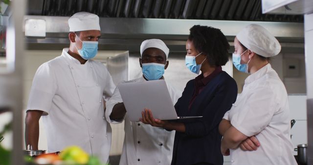 A chef team wearing uniforms and face masks is gathered for a discussion in a commercial kitchen, using a laptop. Ideal for illustrating concepts of teamwork, professional kitchen environments, restaurant management, and hygienic practices in culinary settings.
