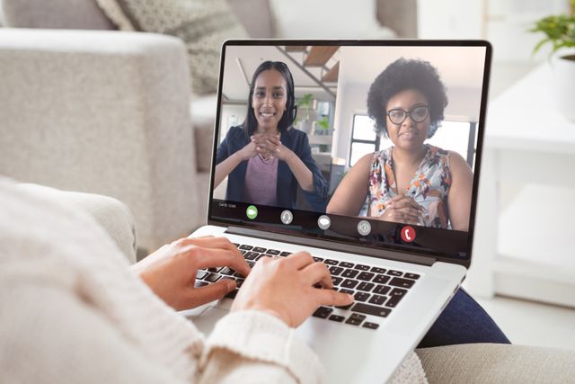 Businesswoman sitting on couch using laptop for video conference with two african american colleagues. Image suitable for articles on remote work, teamwork, virtual meetings, business technology, and work-from-home setups.