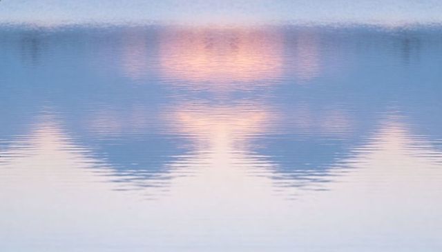 Pastel sunrise reflecting on calm lake with soft ripples and mirrored mountain silhouettes