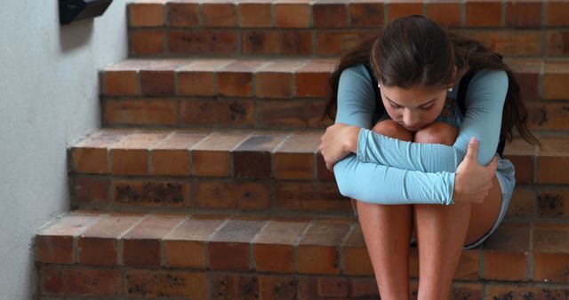 A young Caucasian girl appears distressed as she sits huddled on brick steps, with copy space. Her posture and expression convey a sense of sadness or discomfort, highlighting the importance of mental health awareness.