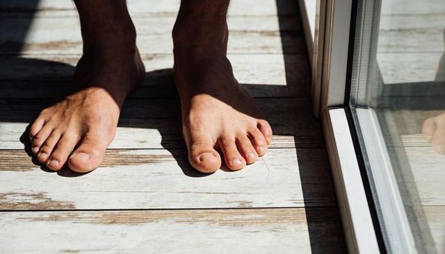 Sunlit bare feet standing on weathered wooden floor by sliding glass door casting reflection