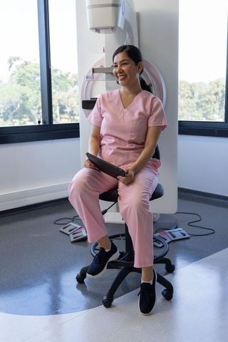 Smiling female mammography technician in pink scrubs holding tablet beside imaging unit