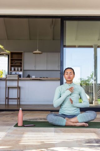 Asian woman practicing mindful meditation on sunlit patio mat by sliding glass kitchen door