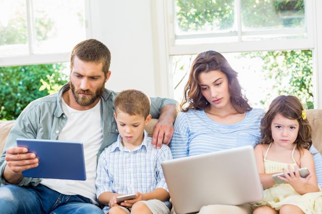 Family sitting on sofa using various digital devices, including a laptop, tablet, and phone. Parents and children are engaged with their gadgets in a living room setting. Ideal for illustrating modern family life, technology use, digital bonding, and home activities.