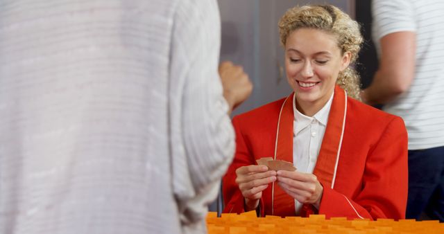 This image shows a woman in an orange jacket smiling while taking part in a card game during a social event. It is perfect for depicting themes of companionship, social gatherings, playfulness, and interactive entertainment during casual events. Ideal for use in advertising social activities, hobbies, and community meet-ups.