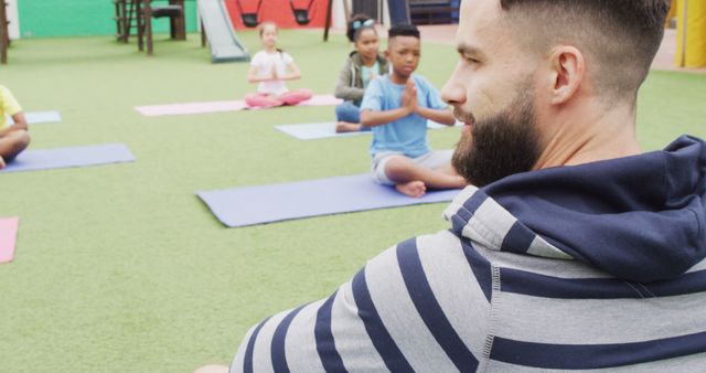Instructor leading an outdoor yoga class with children on mats. Kids focusing on yoga postures and relaxation techniques in a sunny setting. Ideal for promoting children's health and fitness programs, yoga classes, childcare services, summer camps, wellness awareness, and physical education content.