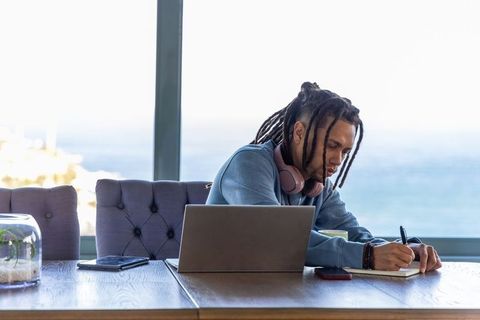 African american man working remotely by ocean window writing in notebook with laptop