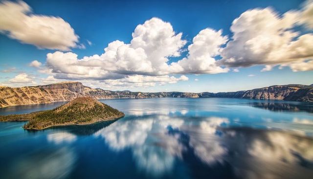Forested conical island reflecting on deep caldera lake with dramatic cloud reflections