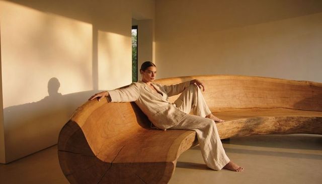 Sunlit minimalist woman lounging on sculpted live-edge wooden bench in linen suit