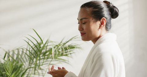 Mid-adult asian woman practicing mindfulness wearing white bathrobe near plants in soft light