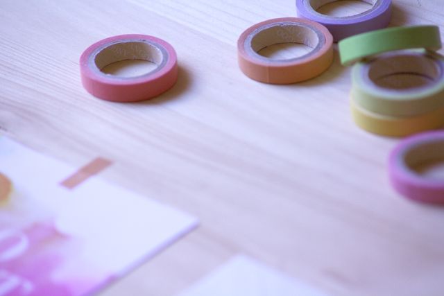 Colorful rolls of washi tape scattered on a wooden surface alongside craft paper. Ideal for themes of DIY crafts, creativity, scrapbooking, and stationary. Perfect for blogs, creative websites, and educational materials about arts and crafts.