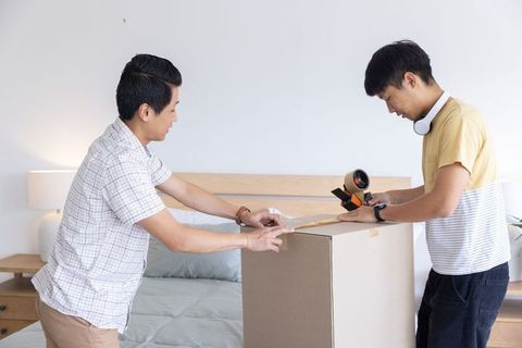 Asian friends packing and sealing moving box in minimalist bedroom, teamwork lifestyle