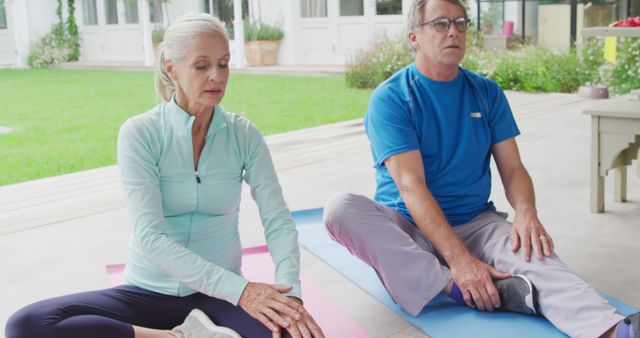 Senior Caucasian couple practices yoga outdoors, with copy space. They're focused on maintaining health and wellness in a serene home setting.