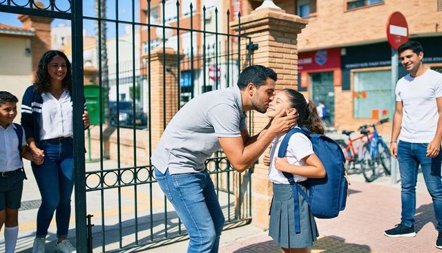 Father kissing daughter at school gate during morning drop-off with backpack and smile