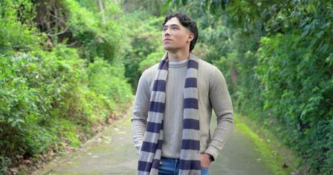 Young man standing on mossy path wearing knit sweater and striped scarf, gazing up