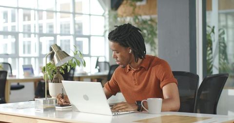 African American man working on laptop and writing in notebook in modern coworking office