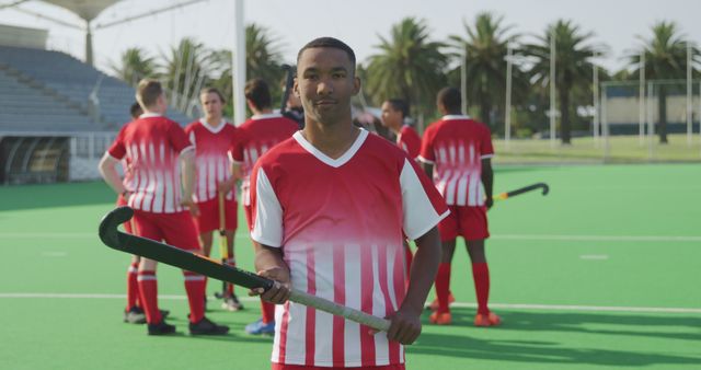 Front view of a confident male field hockey player standing and holding a stick with his teammates in the background on a green outdoor field. This photo could be used for articles about sports team dynamics, advertisements for sports gear, and promotional materials for local field hockey leagues.