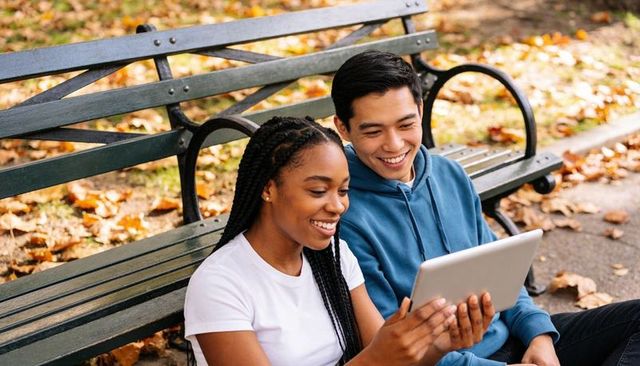 Smiling young couple sharing tablet on park bench surrounded by autumn leaves