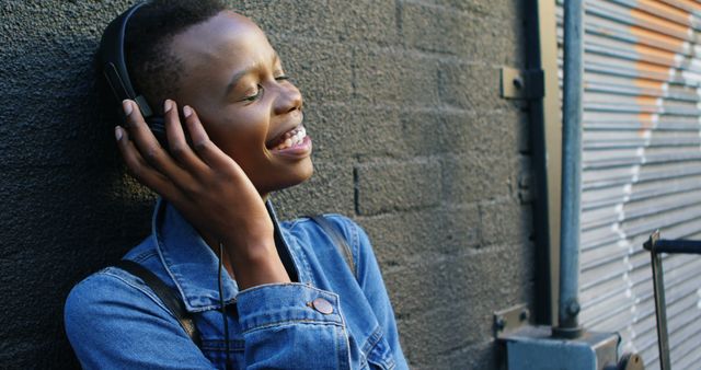 Young woman is enjoying music on headphones while leaning against a brick wall. She is wearing a denim jacket and smiling, appearing joyful and relaxed. Suitable for topics related to music, lifestyle, urban living, youth culture, and summertime relaxation.