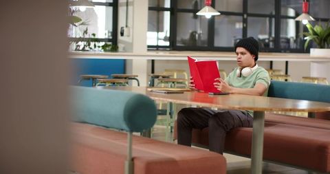 Young man reading red notebook in modern study lounge with headphones at communal table