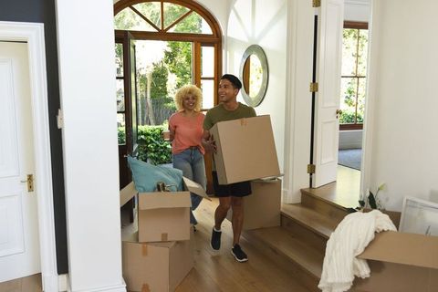 Multiracial couple moving into sunlit home carrying boxes through arched entryway