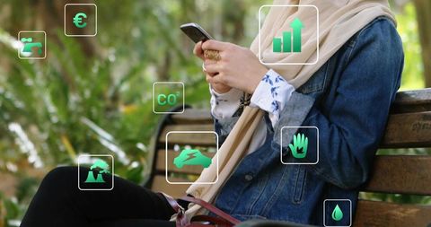Young woman wearing headscarf checking smartphone on park bench with floating eco icons