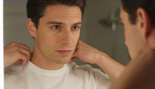Man adjusting white crew-neck shirt in bathroom mirror preparing morning grooming routine