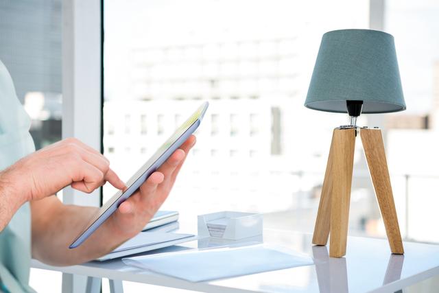 Image depicts a person's hand interacting with a tablet at a desk in a professional setting, with a modern desk lamp in view. Ideal for illustrating technology use in office work, remote working setups, digital workspace concepts, and modern office design.