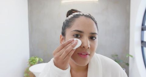 Indian woman cleansing face with cotton pad wearing bathrobe by bathroom window