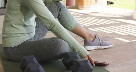 Mid-adult asian woman sitting on patio mat reaching for smartphone near dumbbells