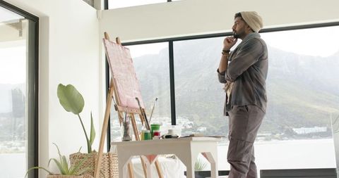 Indian artist standing by window contemplating canvas in minimal sunlit studio overlooking bay