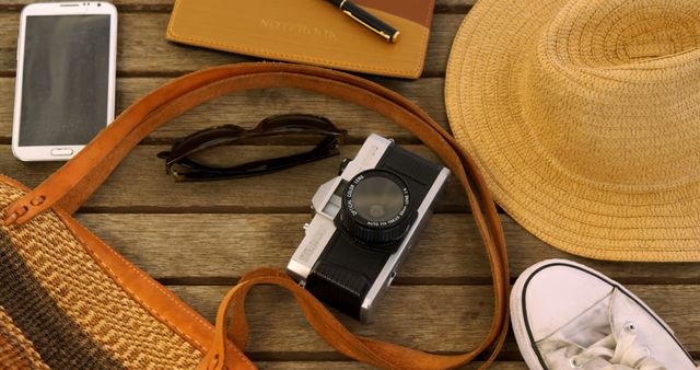 Vintage camera, sunglasses, smartphone, notebook, pen, straw hat, woven bag, and casual shoes on wooden table surface. Perfect for use in travel blogs, adventure advertising, summer holiday promotions, and lifestyle articles. Conveys themes of travel, nostalgia, and preparedness.