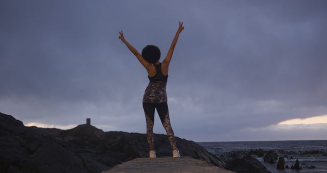Woman standing on rocky shore during early morning, facing ocean with arms raised in celebration, wearing activewear. Ideal for use in health and fitness promotions, outdoor activity advertisements, motivational content, or lifestyle blogs emphasizing nature and achievement.