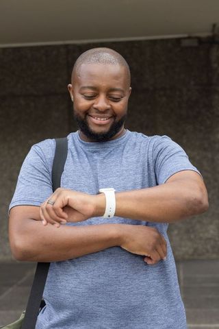 African man checking smartwatch with white band at urban entrance, smiling and wearing crossbody str