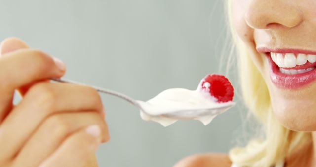 A close-up of a young Caucasian woman about to eat yogurt with a raspberry on a spoon, with copy space. Her smile suggests enjoyment of the simple pleasure of eating a healthy snack.