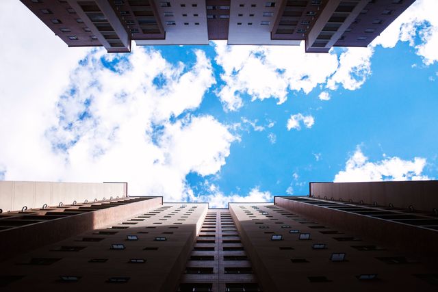 Urban scene with tall buildings framing cloudy blue sky in city. Interesting perspective and symmetry make this suitable for architectural design, city planning, or motivational themes. Can be used in brochures, presentations, or websites discussing urban living, real estate, or travel.