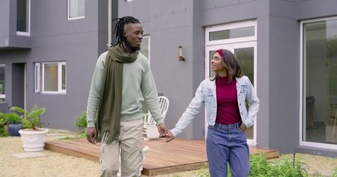 Diverse Couple Walking Hand-in-Hand Beside Modern Gray Home with Wooden Deck