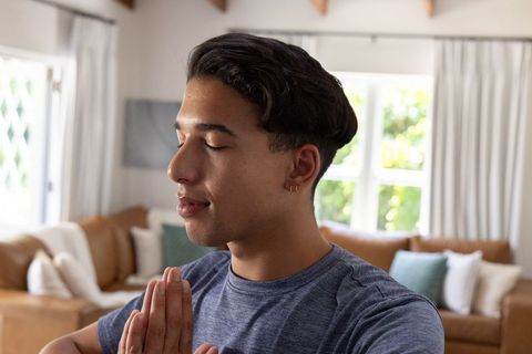 Young man meditating with palms together in sunlit living room calm home wellness portrait