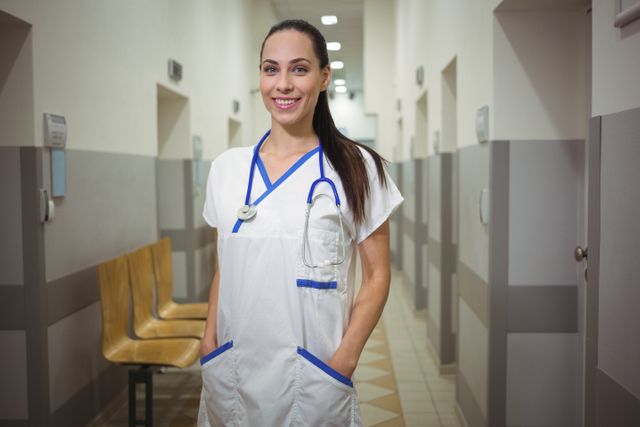 Confident female nurse standing in a hospital corridor with a stethoscope around her neck and hands in her pockets, embodying professionalism and compassion. Ideal for use in healthcare articles, medical advertisements, hospital brochures, and educational materials.