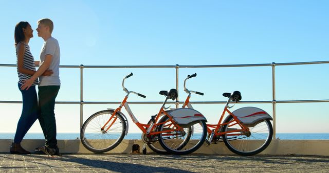 A young Caucasian couple enjoys a romantic moment by the sea, with two orange bicycles resting beside them, with copy space. Their affectionate embrace captures a leisurely day spent cycling and relaxing by the waterfront.