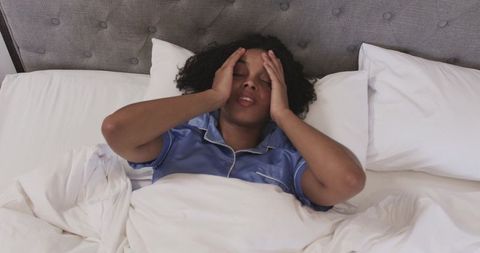 African american woman pressing temples while lying in bed in blue satin pajamas