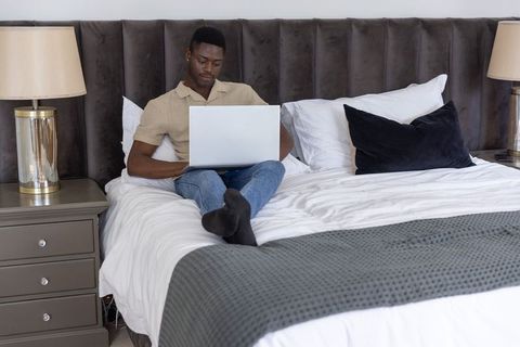 African american man working on laptop in modern neutral bedroom with tufted headboard