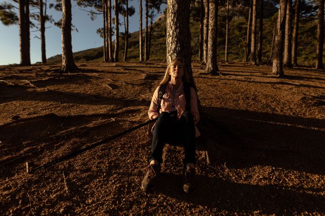 Senior woman enjoying a peaceful moment during a mountain hike, sitting against a tree with her eyes closed. Ideal for use in articles or advertisements related to outdoor activities, healthy aging, retirement lifestyle, and nature retreats.