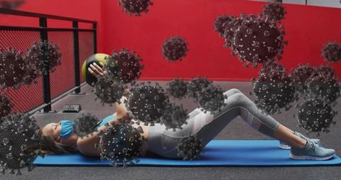Masked woman exercising with medicine ball on mat amid floating virus illustration overlay