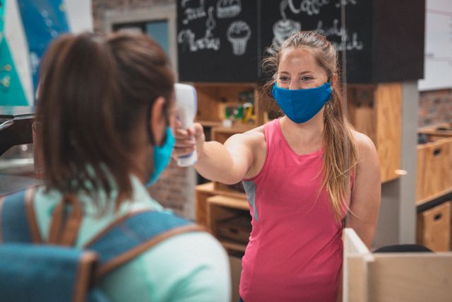 Caucasian female gym receptionist waring face mask taking temperature of female client. fitness and leisure during coronavirus covid 19 pandemic.