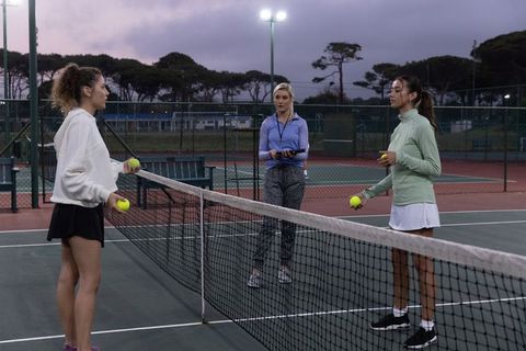 Evening tennis coaching session with diverse players and coach timing ball drill on court