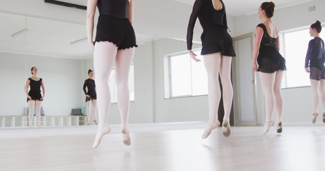 Ballet dancers jumping in unison during practice in a spacious, well-lit dance studio. Ballet practice clothing and focused expressions indicate a serious training session. Ideal for articles on performing arts, dance education materials, promotional images for dance schools, and fitness-related posts.