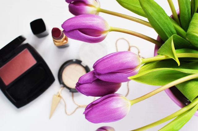 Purple tulips arranged next to various makeup products, including compact powder, lipstick, and blush, on white background. Ideal for beauty blogs, makeup tutorials, lifestyle websites, and floral arrangement articles due to its vibrant colors and appealing composition.
